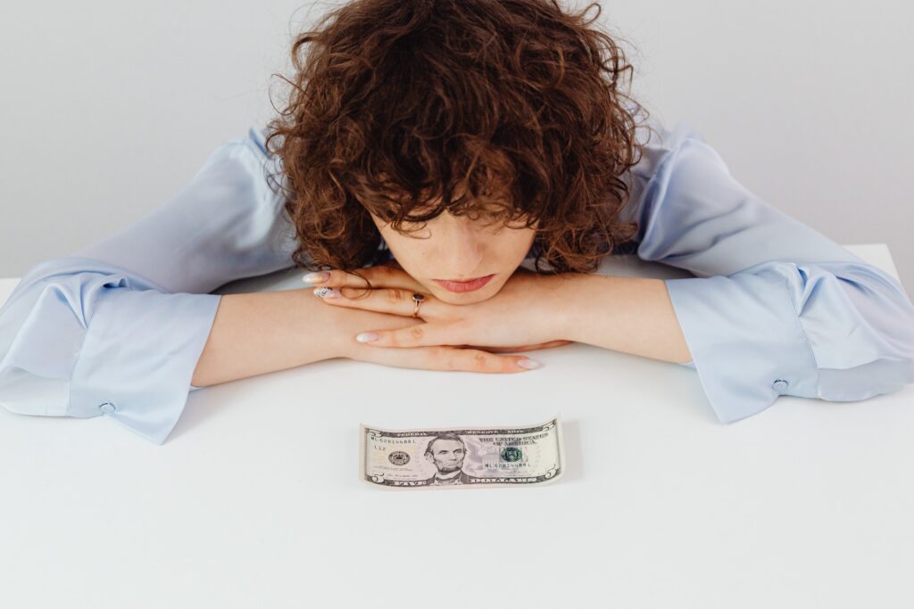 A woman with brown curly hair rests her head on her hands while she stares at a five dollar bill.