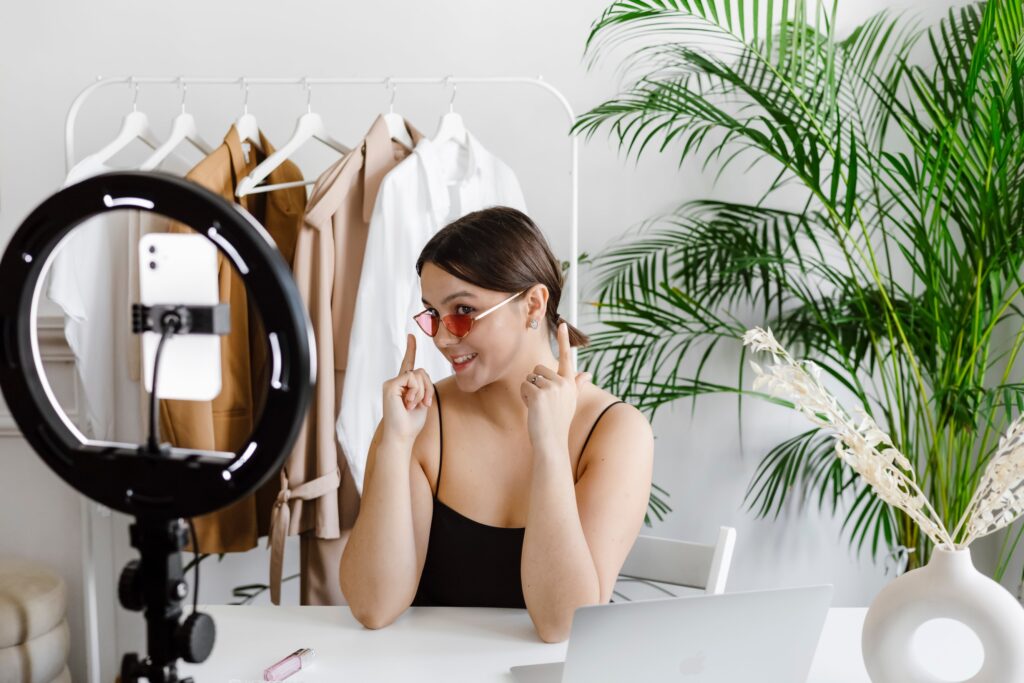 A young, female influencer in front of a ring light showing off sunglasses during a broadcast on her cell phone 