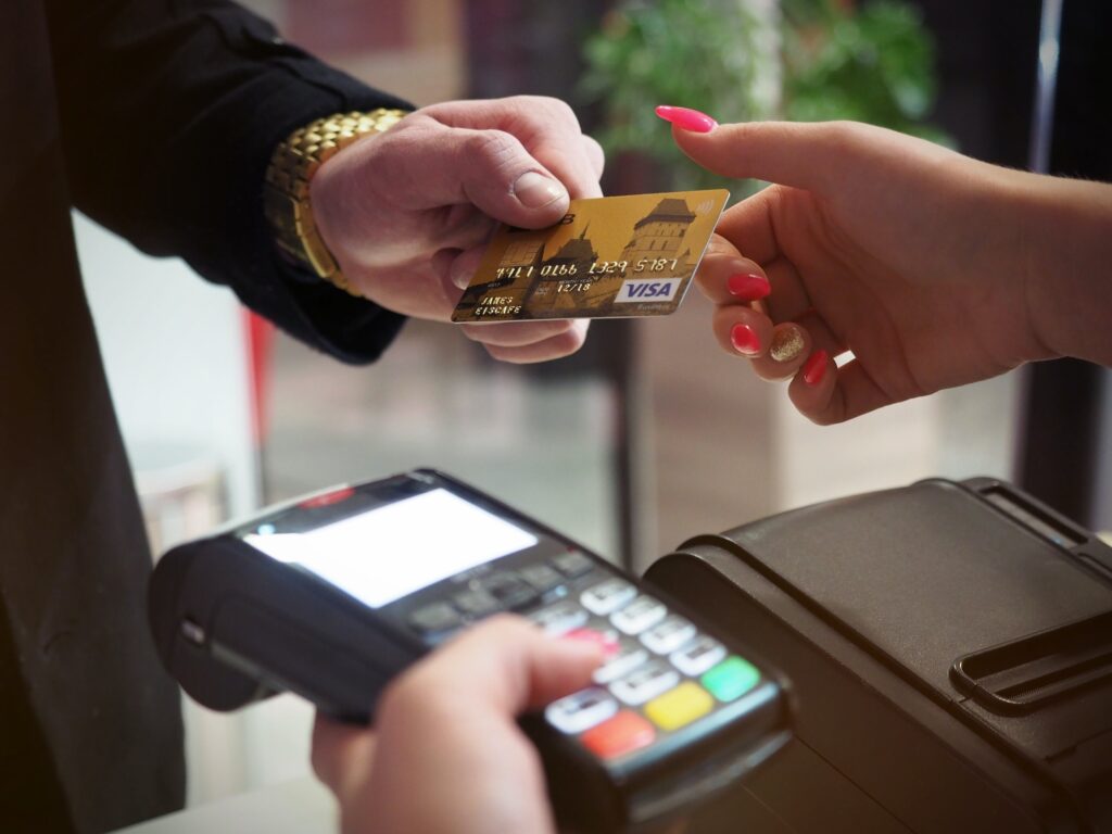 A man wearing a black business suit and a gold watch hannding a gold VISA card to a female merchant with bright pink fingernails