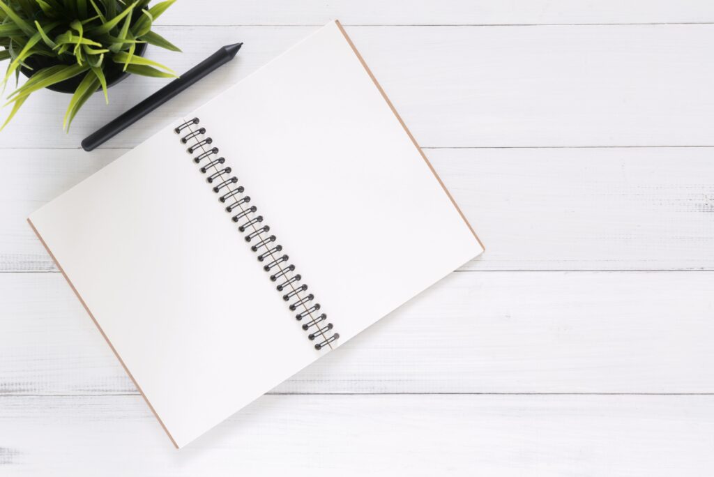 A top down view of a white table with a potted green plant in the top left corner, a pen, and a blank, open notebook