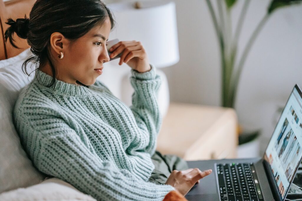 A young, dark haired woman sitting on a couch wearing a light blue sweater looking at a laptop shopping with a credit card in her left hand