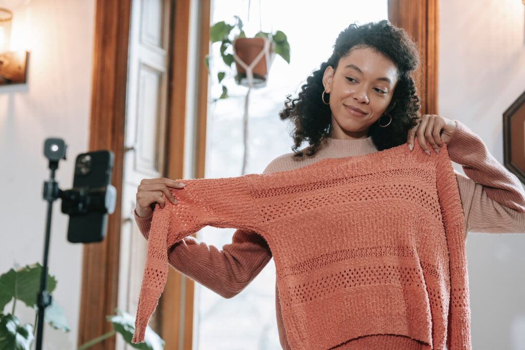 A young dark haired woman showing off a peach colored sweater in front of a livestreaming smartphone set up