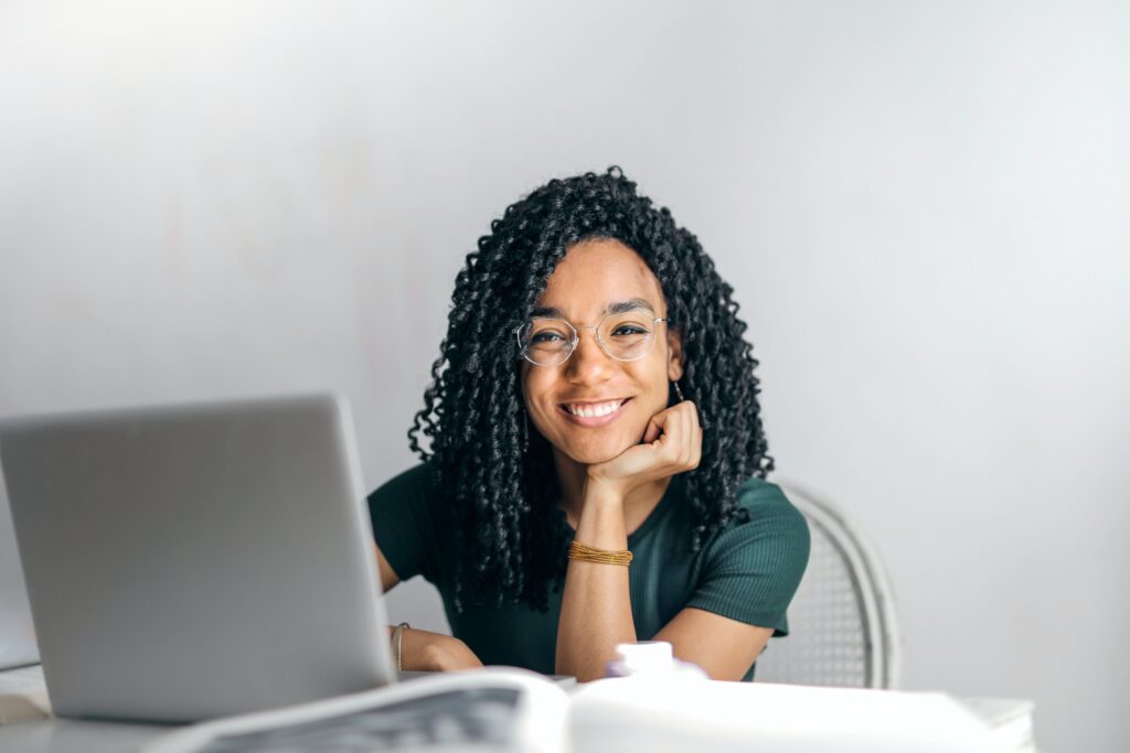 A young dark haired woman sitting at a table, in fron of an open laptop posed with her open hand uner her chin looking at the camera