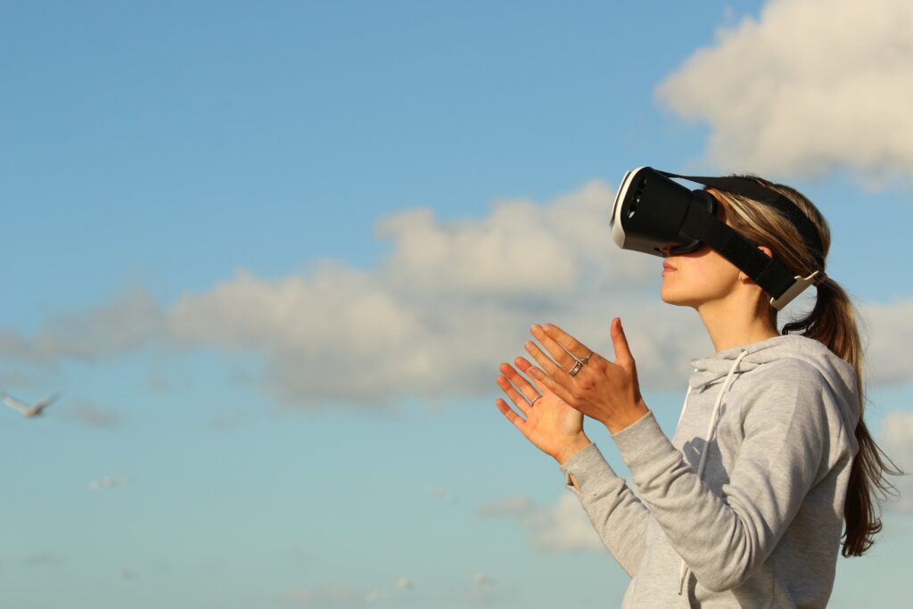 A young, blonde woman looking through virtual googles outside with her hands outraised 