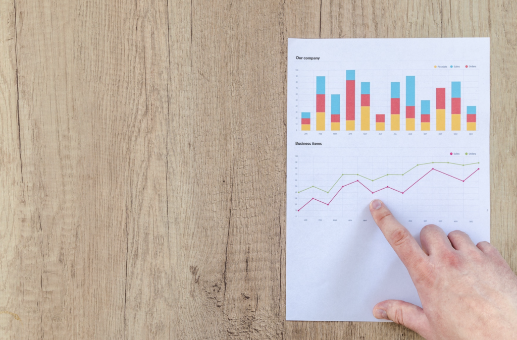 a top down view of a light brown wooden table, a white sheet of paper showing upwardly trending data and a single right hand pointing to the upward trending graph with an index finger